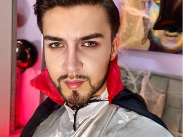 DaltonWhite, avec des yeux verts, des cheveux foncés et une barbe, portant une chemise argentée brillante avec un col rouge et une cape noire, regardant la caméra avec un léger sourire..