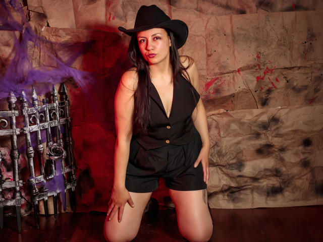MargarettCooper, kneeling, wears a black cowboy hat, sleeveless black vest, and shorts, with long brown hair, and a confident expression, showing her tits and ass.