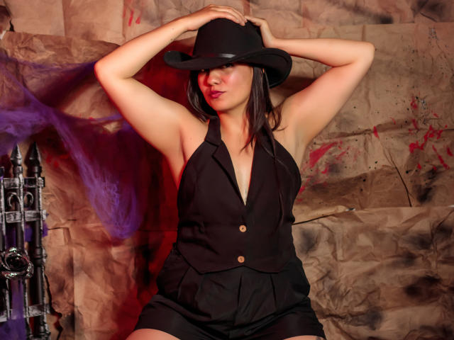 MargarettCooper with arms up, wearing a black hat, sleeveless black vest, and shorts, showing her tits and ass, against a textured brown background with purple smoke.