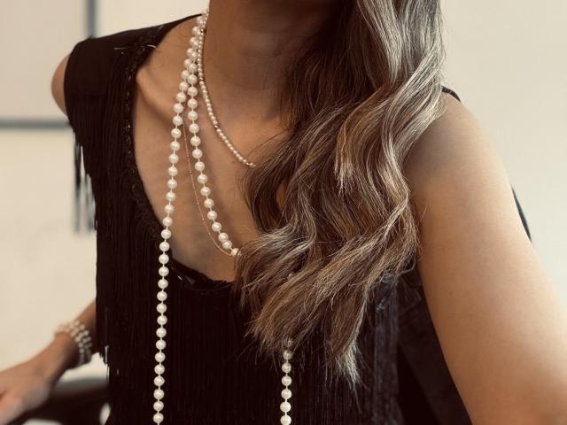 AdharaCooperr's upper body, wearing a black fringe dress, with a long, wavy brown hair. She has a double-strand pearl necklace, the pearls are white and shiny. The dress has a deep V-neck, and the fringe adds texture. Her right arm is slightly out of frame, showing a simple bracelet. The image focuses on her neck and chest area. The lighting is soft, highlighting the pearls and dress.