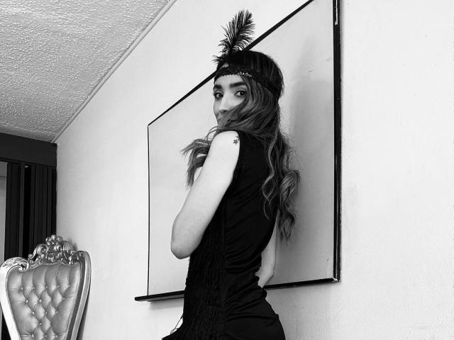 AdharaCooperr with long wavy hair, wearing a black fringed top, black headband with feather, looking over shoulder, small tattoo on upper arm, black-and-white photo.