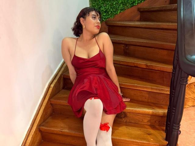 NoemiThornee, with curly brown hair, sits on wooden stairs in a red dress, showing her tits, and white thigh-highs with a red bow, looking to the side.