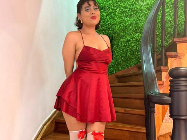 NoemiThornee, avec des seins de taille moyenne, des hanches rebondies, portant une robe en satin rouge, des bas blancs montants jusqu'à la cuisse avec des nœuds rouges, debout près d'un escalier en bois, un mur vert en arrière-plan..
