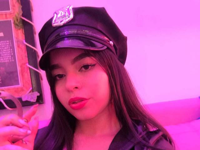 LissyCarter with dark hair, wearing a black police hat, holding a pen, pinkish-purple lighting, red lipstick, slightly parted pink lips, looking at camera, pinkish-purple skin tone.