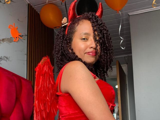 ArtemisaS with curly black hair, red devil horns, and red wings, wearing a red dress, smiling at the camera, showing her cute brown skin and red lips.