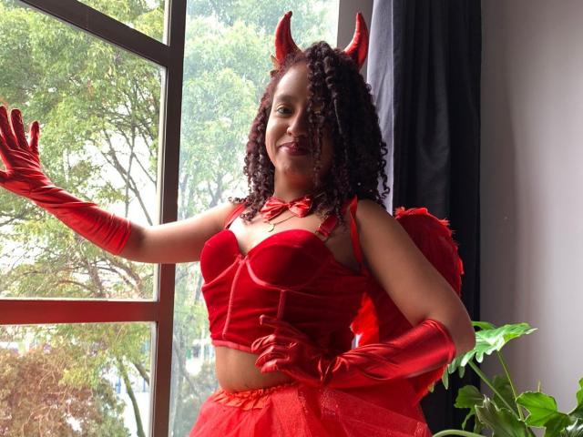 ArtemisaS with curly brown hair, red devil horns, red velvet bustier, red gloves, and red tulle skirt, smiling, standing by a window, showing her red-boobed tits.