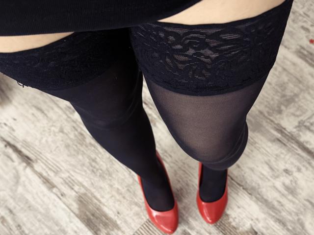 Close-up of Rubbi's lower body. Rubbi wears sheer black stockings with lace tops, slightly visible under a black skirt. Rubbi's feet are in red pointed shoes. The lace texture contrasts with the sheer material of the stockings. The image focuses on the lower half, with a slight upward angle. The wooden floor has a greyish texture, adding to the visual contrast. The composition emphasizes the interplay between the lace, sheer material, and the bold red shoes.