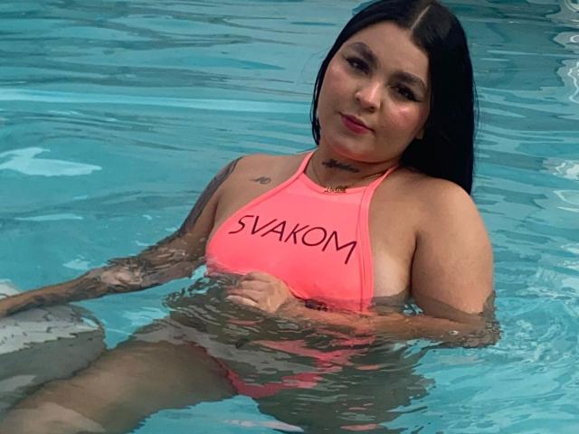 MelodyBlossom with dark hair, pink swim top, and tattoos, sitting in a pool, wearing a neon pink "SVAXOM" top, holding her tits, with a subtle smile.