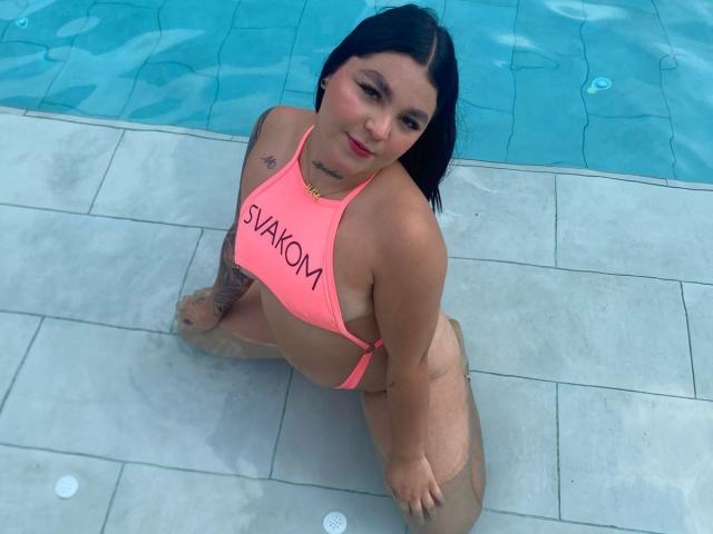 MelodyBlossom, con i capelli neri, un bikini rosa e la scritta "STAKOM" sulla parte superiore, è inginocchiata in una piscina, mostrando il seno e un tatuaggio sul braccio destro..
