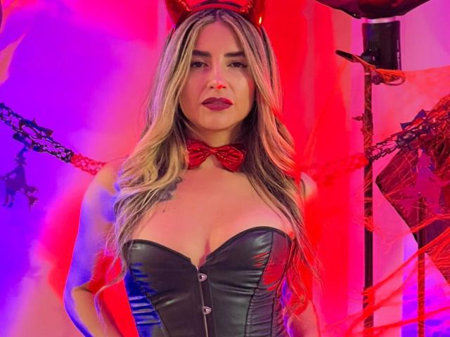 SofyaLane with long blonde hair, red devil horns, and a black leather corset, showcasing her big tits, red bowtie, and red gloves, under colorful red and purple lighting.