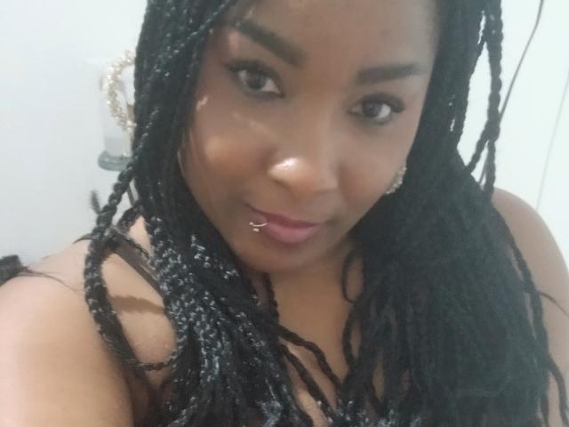 DakotaScoth with dark braided hair, brown eyes, and a lip piercing, smiling slightly, showing off her shiny nose and wearing a black strap top.