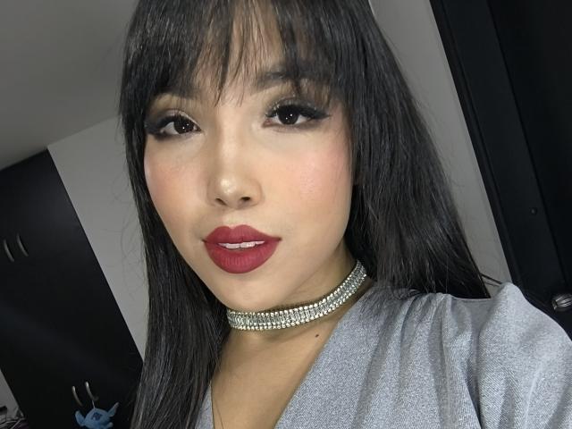 Heyly with straight black hair, red lipstick, and sparkling choker, looking directly at the camera, wearing a gray shirt.