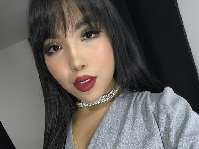 Heyly's got her face close to the camera, lips painted deep red, wearing a sparkly choker, black hair, and a grey top.