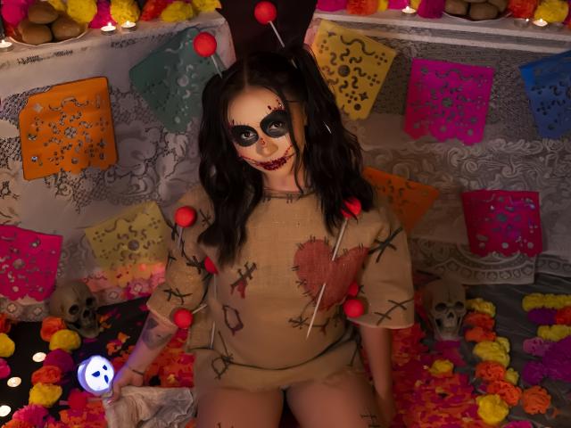 AilenTovar with dark hair, face painted like a Day of the Dead skull, wearing a beige dress with red stitches, and red balls, sitting on colorful flowers.