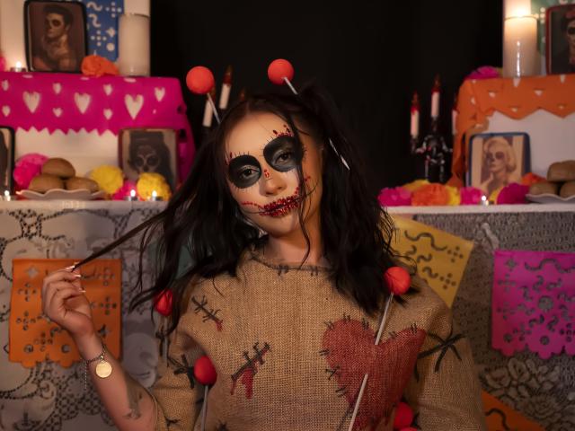 AilenTovar with dark makeup, bloody red mouth, and black hair pigtails, wearing a burlap shirt with red heart and stitches, holding a stick with red balls.