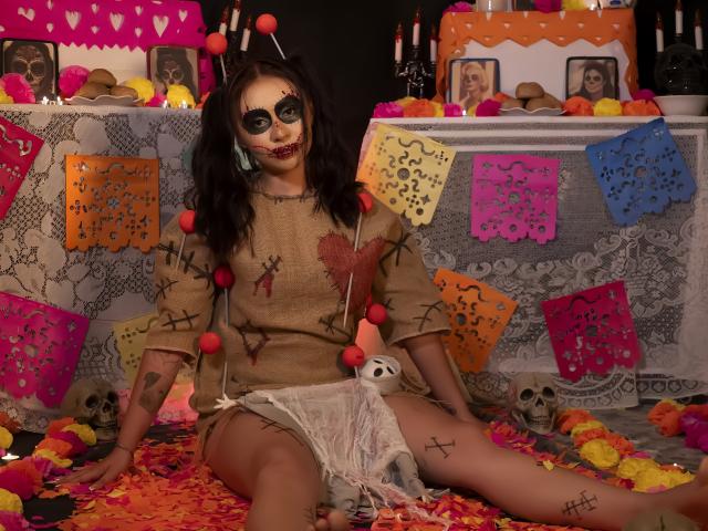 AilenTovar with dark makeup, bloody teeth, and burlap dress sits on colorful flowers, surrounded by skulls and paper decorations. She's wearing a white cloth over her pussy.