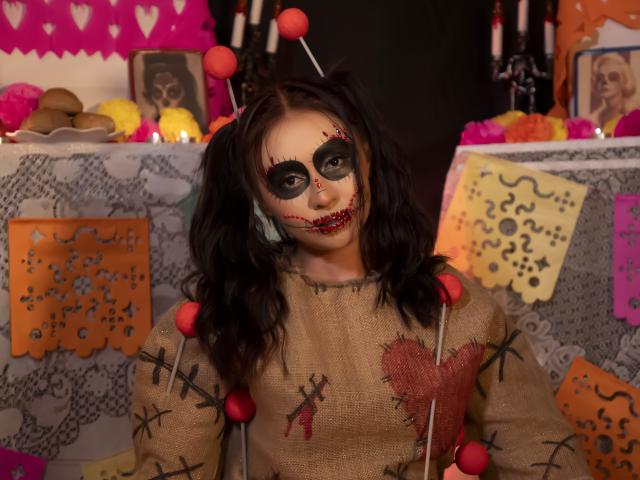 AilenTovar with black pigtails, white face paint, red lipstick, and black eye makeup, wearing a burlap shirt with red heart and stitch designs, and red pom-poms.