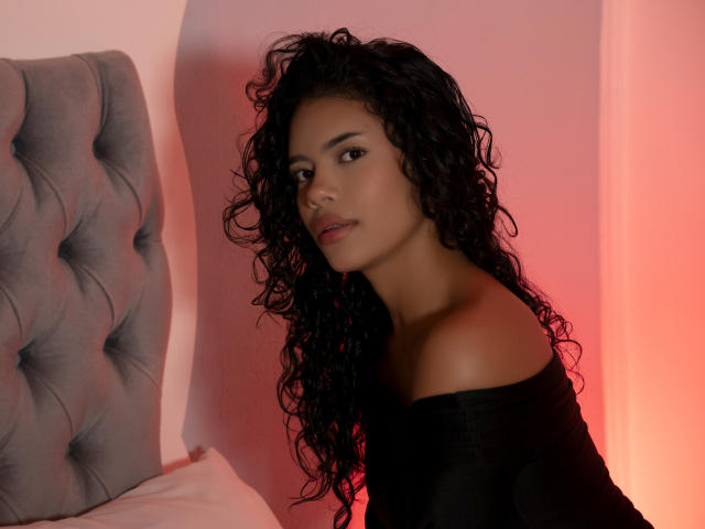 ZoeRios with curly black hair, off-shoulder black top, and sultry expression, sitting by a tufted grey headboard, glowing pink light in the background.