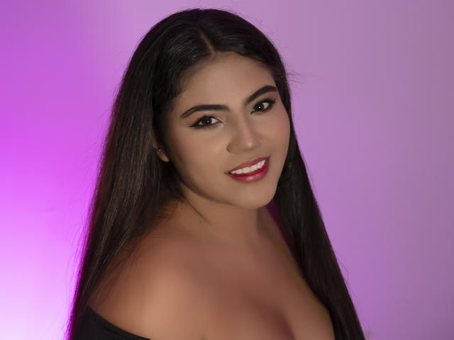 VictoriaRosel with long dark hair, red lipstick, and black off-shoulder top, showing her tits and smiling at the camera. She has a cute face and purple gradient background.