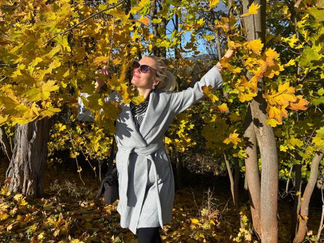LadyMargaret-hot with blonde hair, wearing a grey coat, black tights, and sunglasses, stands in a yellow-leaved forest, arms raised, touching branches, smiling. Autumn sunlight.