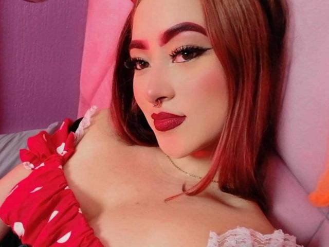 SamaraCuttie está usando uma blusa vermelha com bolinhas, que mostra seus seios, com maquiagem incluindo delineador escuro e batom vermelho, e um piercing no nariz, deitada em almofadas rosa e roxas..