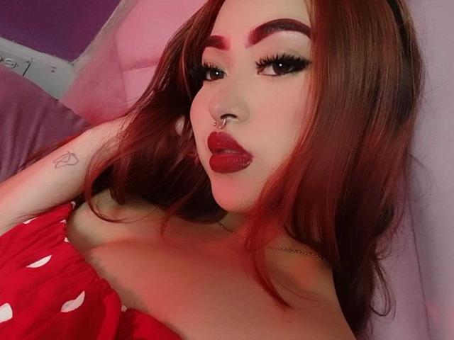 Close-up of SamaraCuttie with long, reddish-brown hair and bold, dark eyebrows. She has thick, black eyeliner and long, dramatic lashes. Her nose is pierced with a small ring. She wears bright red lipstick and a red, strapless top with white polka dots. A small heart tattoo is visible on her left arm. The image has a soft, pinkish light, emphasizing her makeup and top. The composition is tight, focusing on her face and upper chest.