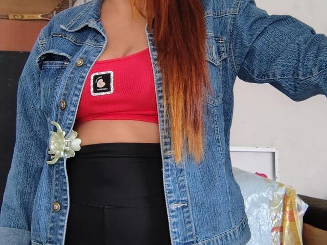 SammantaHott con cabello rojo largo, chaqueta vaquera, top deportivo rojo que muestra sus pechos, pantalones negros de cintura alta y un accesorio con una flor verde en la chaqueta..