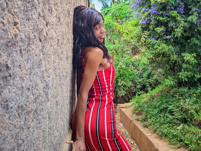 VictorineStone with long black hair, wearing a red and black striped dress, leaning against a rough wall, showing her ass and smiling, surrounded by greenery and purple flowers.