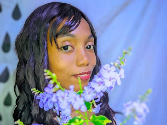 VictorineStone with wavy black hair, brown skin, and pink lipstick, holding purple and white flowers in front of her face, smiling softly. Her tits are partially visible.