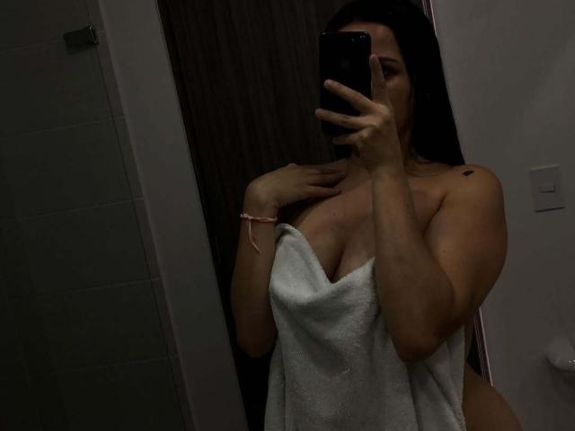 MiaAylen with dark hair, holding phone, covering boobs with white towel, right hand on chest, pink bracelet, black tattoo on right shoulder, dimly lit bathroom mirror selfie.