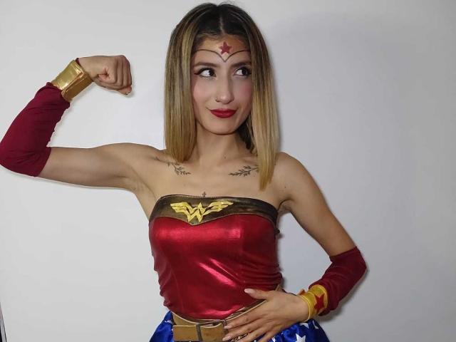 Kaiix, mit blonden Haaren, einem roten, trägerlosen Wonder Woman-Top, einer goldenen Tiara, einem angehobenen Arm, roten Armwärmern und einem blauen Rock, hat kleine Brüste, Tätowierungen auf der Brust und roten Lippenstift..