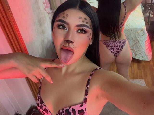MaddisonCarter with long black hair, leopard print bra and thong, sticking out tongue, finger near mouth, face paint spots, mirror reflection showing her ass, in dimly lit room.