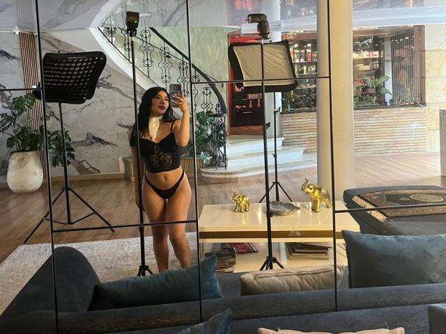 MaddisonCarter, with long black hair, stands in black lace bra and panties, taking a mirror selfie in a modern living room, surrounded by photography equipment.
