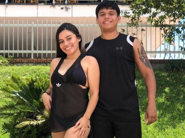 PrettyHotCouple69 with long black hair and small boobs in a sheer black top, stands next to a tall, tattooed guy in a black Under Armour shirt. Both smiling.