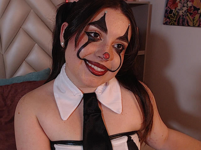 VicluX has dark hair in pigtails, black eye makeup, red nose, and white collar with black tie, smiling at the camera, showing freckles and red lips.
