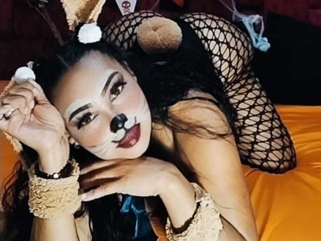 MelanyContrera in a black fishnet bodysuit, lying on an orange bed, with bunny ears and a black nose on her face, her ass up, hands near her mouth.