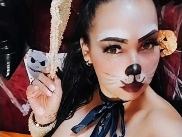 MelanyContrera with dark makeup, black nose, and red lipstick, holding a rope, wearing a black ribbon choker, and a black sleeve, with a Jack Skellington pillow.