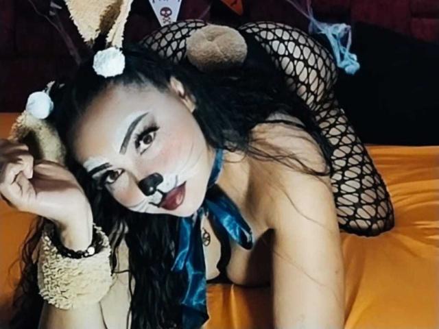 MelanyContrera with black fishnet stockings, bunny ears, white makeup, and dark lipstick, lying on an orange bed, showing her tits and ass, wearing a blue ribbon.