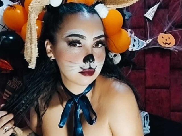 MelanyContrera with dark hair in a ponytail, cat ears, white face paint, black nose, red lipstick, and black ribbon, topless, showing her tits, in a Halloween-themed shot.