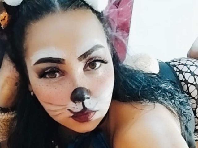 MelanyContrera with dark makeup, black nose, and red lipstick, wearing a black fishnet top, lying down, looking at the camera with a sexy, playful expression.