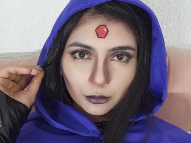 JeneKurz has dark purple lipstick, a red hexagon tattoo on her forehead, and a blue hoodie. She's looking at the camera with brown eyes, black eyebrows, and long black hair.