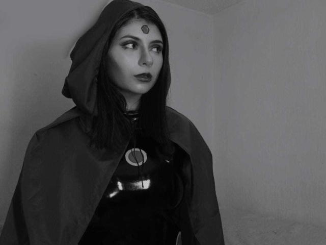 Black-and-white photograph of JeneKurz. JeneKurz has dark, straight hair, and a forehead mark resembling a small heart. She wears a dark hooded cloak over a shiny, black latex dress with a circular emblem on the chest. JeneKurz's expression is serious, looking to the right. The grayscale contrast highlights her sharp features and the texture of the cloak. The image has a minimalist, moody vibe.