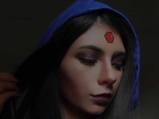 JeneKurz has dark, straight hair, wearing a blue hood, with a red hexagon on her forehead. She's got purple eyelids and dark lipstick, looking down.