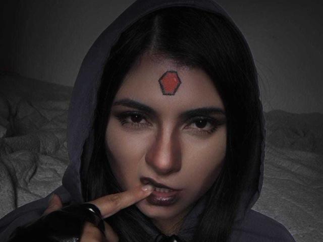 JeneKurz with dark hair, hooded, red hexagon tattoo on forehead, finger on slightly open mouth, dark eyes, and subtle makeup, in dim lighting.