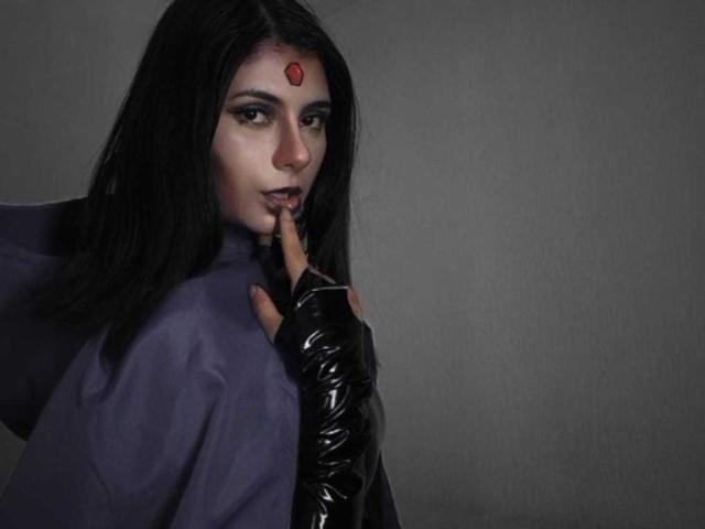 JeneKurz with dark hair, red forehead jewel, purple robe, black glossy fingerless gloves, finger in mouth, looking seductively over shoulder, grey background.