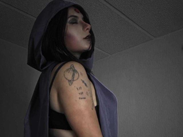 JeneKurz with black hair, hooded purple cape, black bra, tattoos on right arm, dark lipstick, eyes closed, in dimly lit room.
