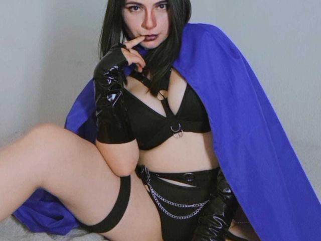 JeneKurz with black hair, wearing a blue cape, black bra, and shiny black gloves, touching her finger to her lips, sitting with one leg up, showing her thighs.