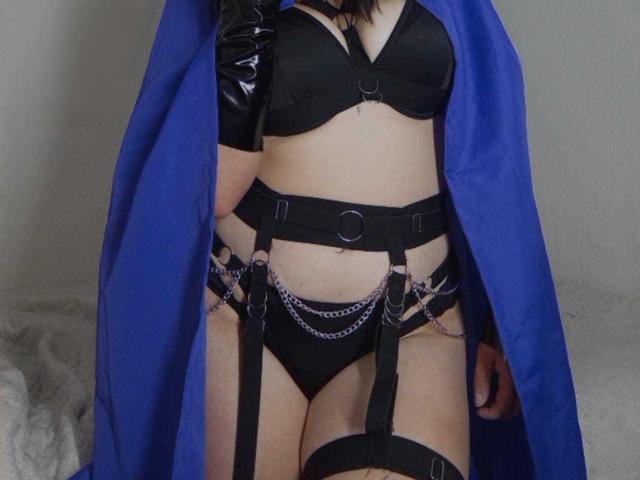JeneKurz in black lingerie with straps, chains, and a blue cape, wearing black latex gloves, showing her ass and tits, standing with her legs together.