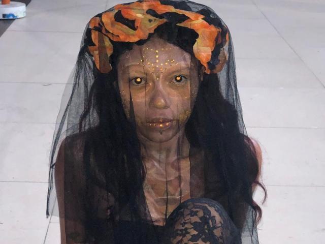 LunaSweets a la peau foncée, les yeux jaunes et les cheveux noirs. Elle porte un voile noir orné de fleurs orange et de points dorés sur son visage, et est assise, le genou recouvert de dentelle..