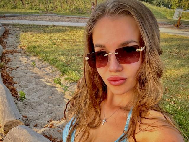 LightTeresa mit welligem, braunem Haar, die dunkle Sonnenbrillen mit goldenen Details und ein blaues Bikini-Oberteil trägt, hat eine Kreuzkette um den Hals und leicht geschminkte, rosafarbene Lippen. Im Hintergrund ist eine sonnendurchflutete, grüne Wiese zu sehen..